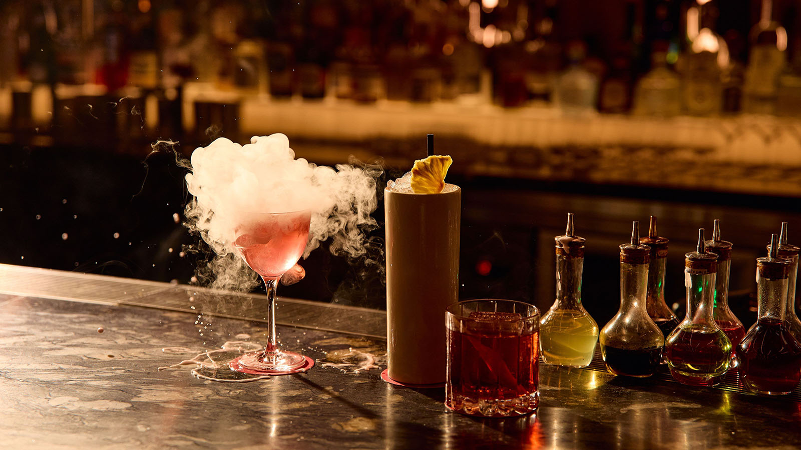 Three cocktails on a dimly lit bar counter with one smoking pink drink, a tall pineapple cocktail and a classic dark spirit glass surrounded by bitters bottles.