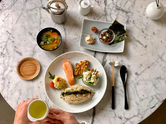Traditional Japanese breakfast set (Asagohan) served on a white marble table, featuring rice, salmon, tamagoyaki, miso soup, and green tea.