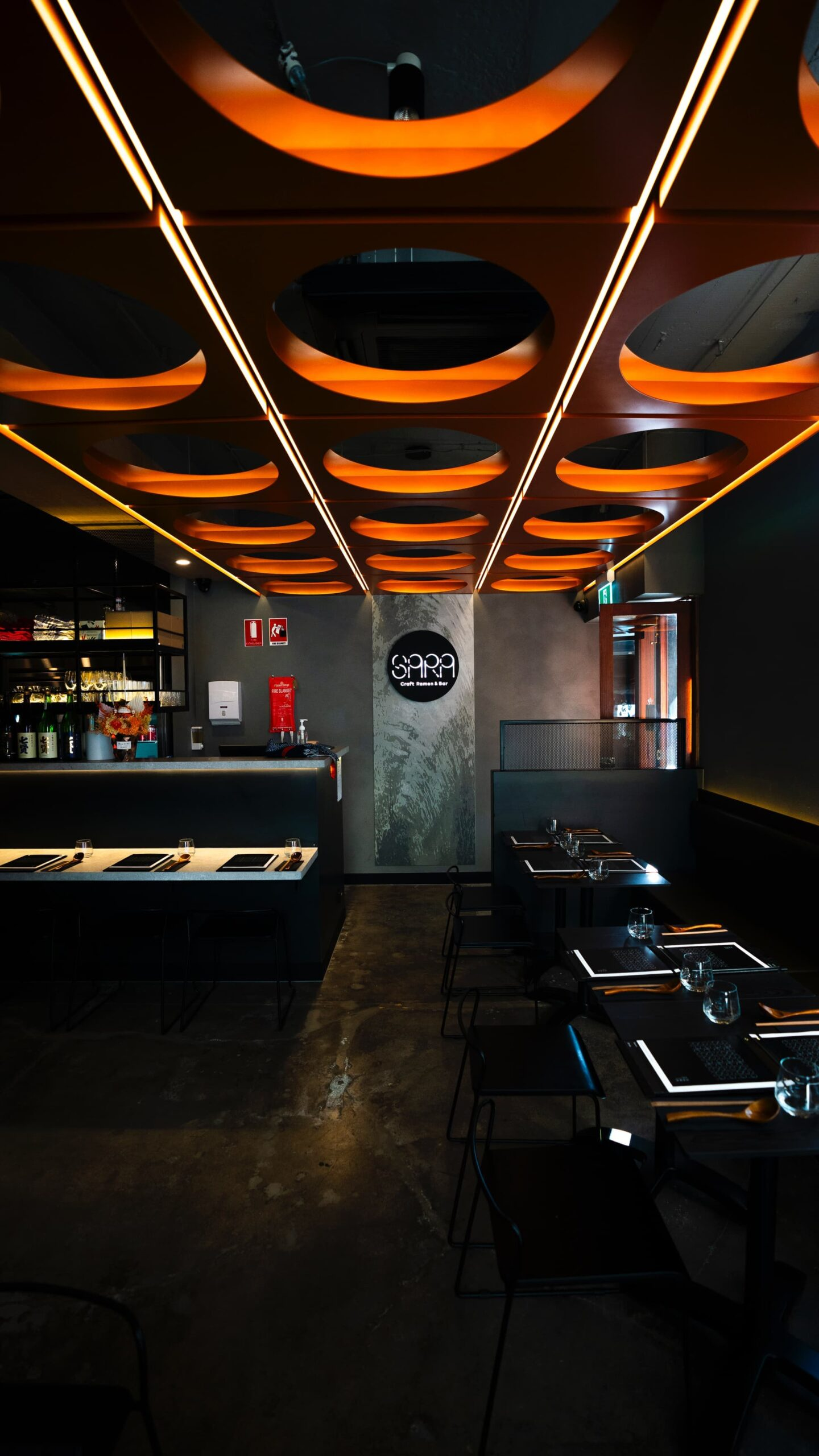 Modern, dimly-lit interior of Sara Craft Ramen Bar with dark seating and circular orange ceiling lighting.