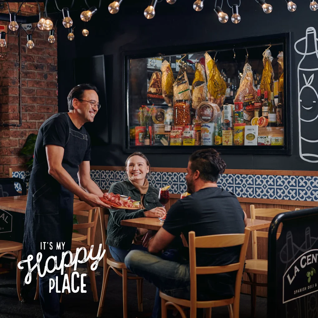 Two customers smiling while seated outside La Central Deli as a worker serves them a dish of food