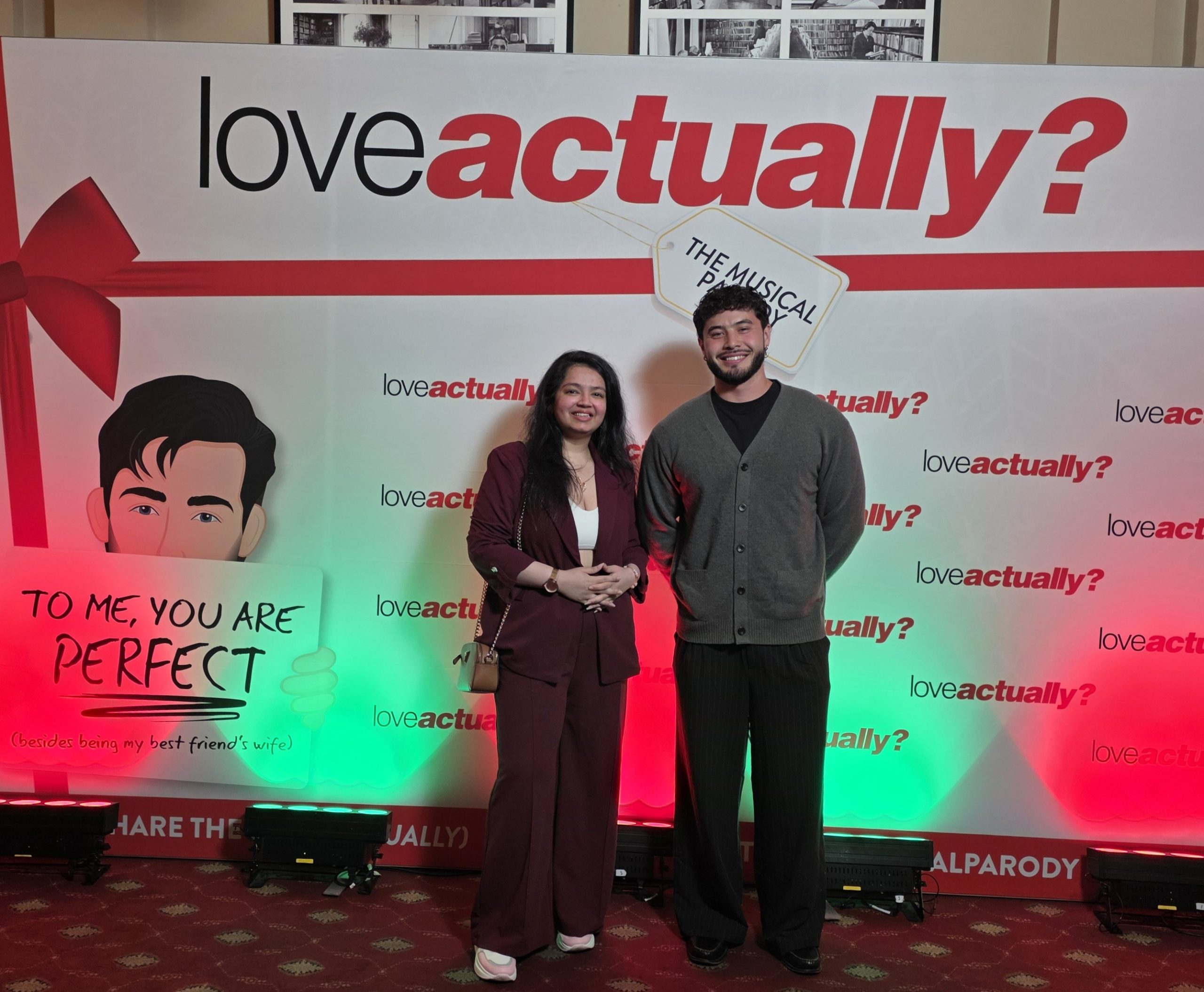 The cast of Love Actually? The Musical Parody holding playbills on stage in front of the red doors set.