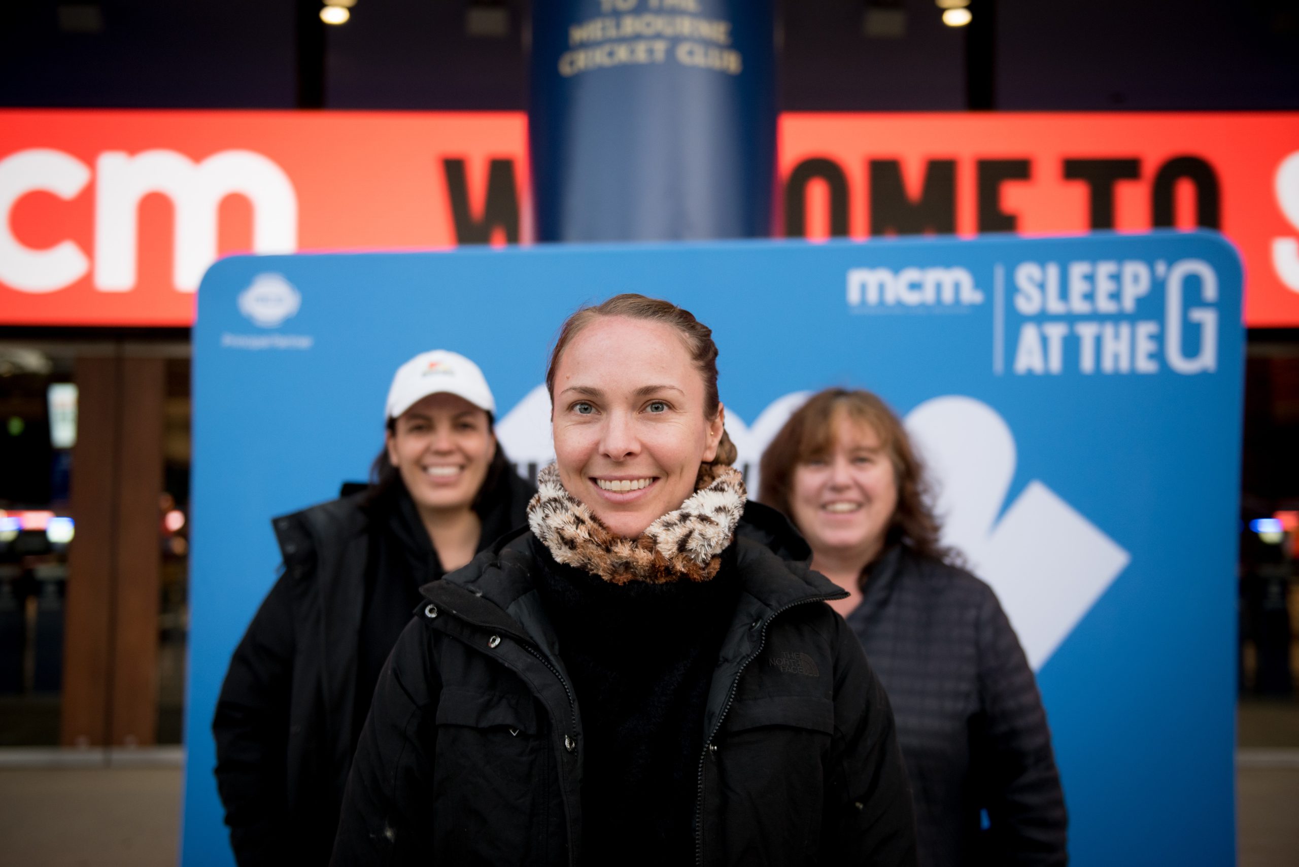 Sleep at the G 2026 participants smiling in front of Melbourne City Mission Sleep at the G sign at Melbourne Cricket Ground entrance