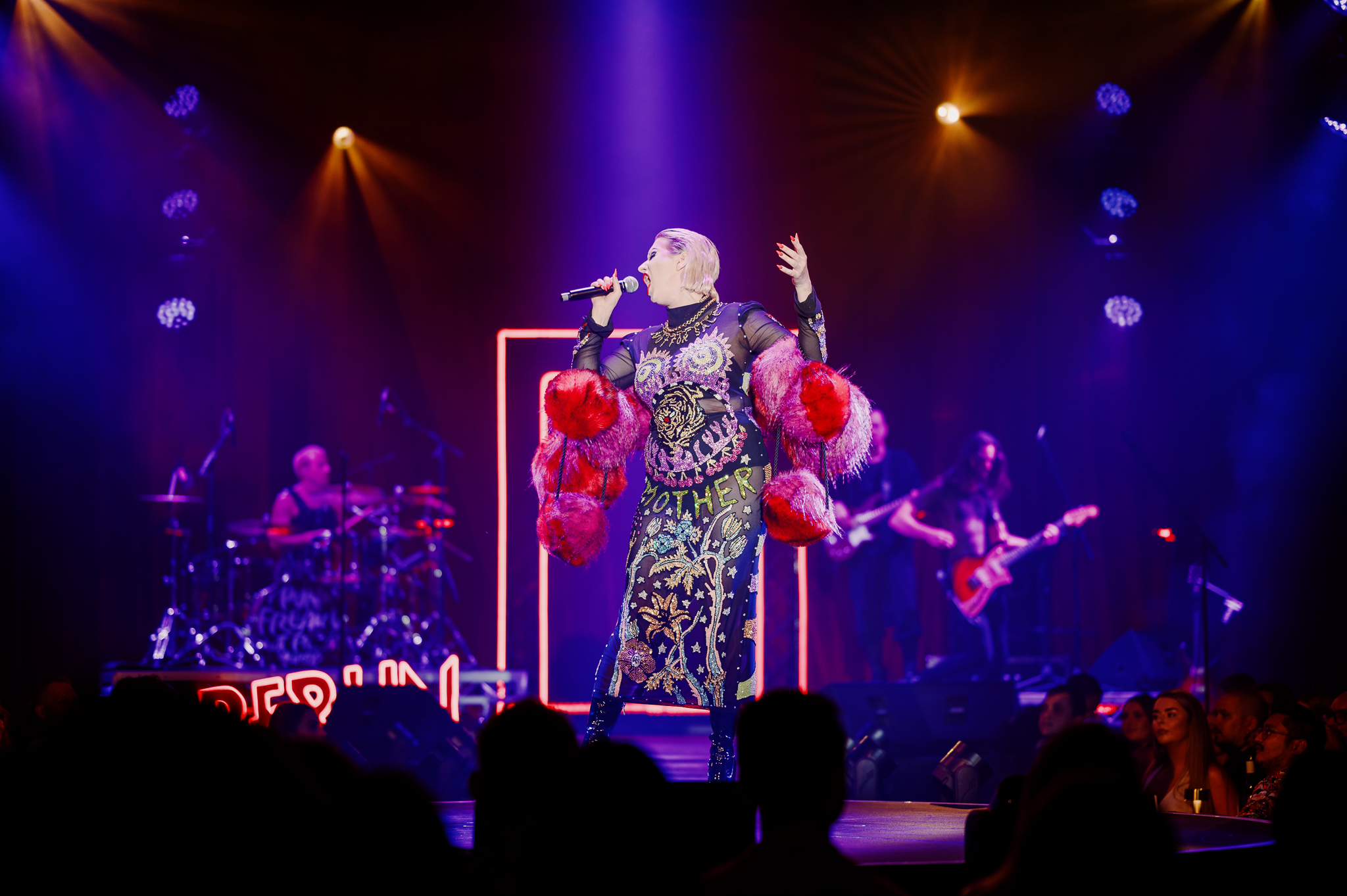 Bernie Dieter singing on stage at Club Kabarett in a sequined costume adorned with red fur pom poms under blue and red lighting