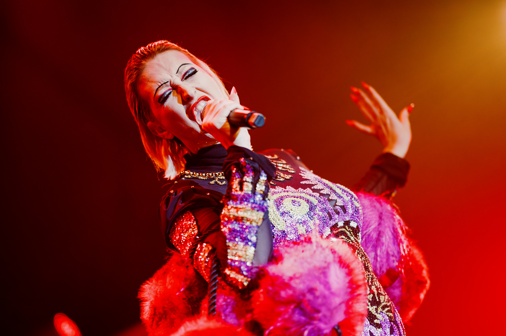 Close up of Bernie Dieter singing passionately on stage under red lighting in a sequined costume with pink fur trim