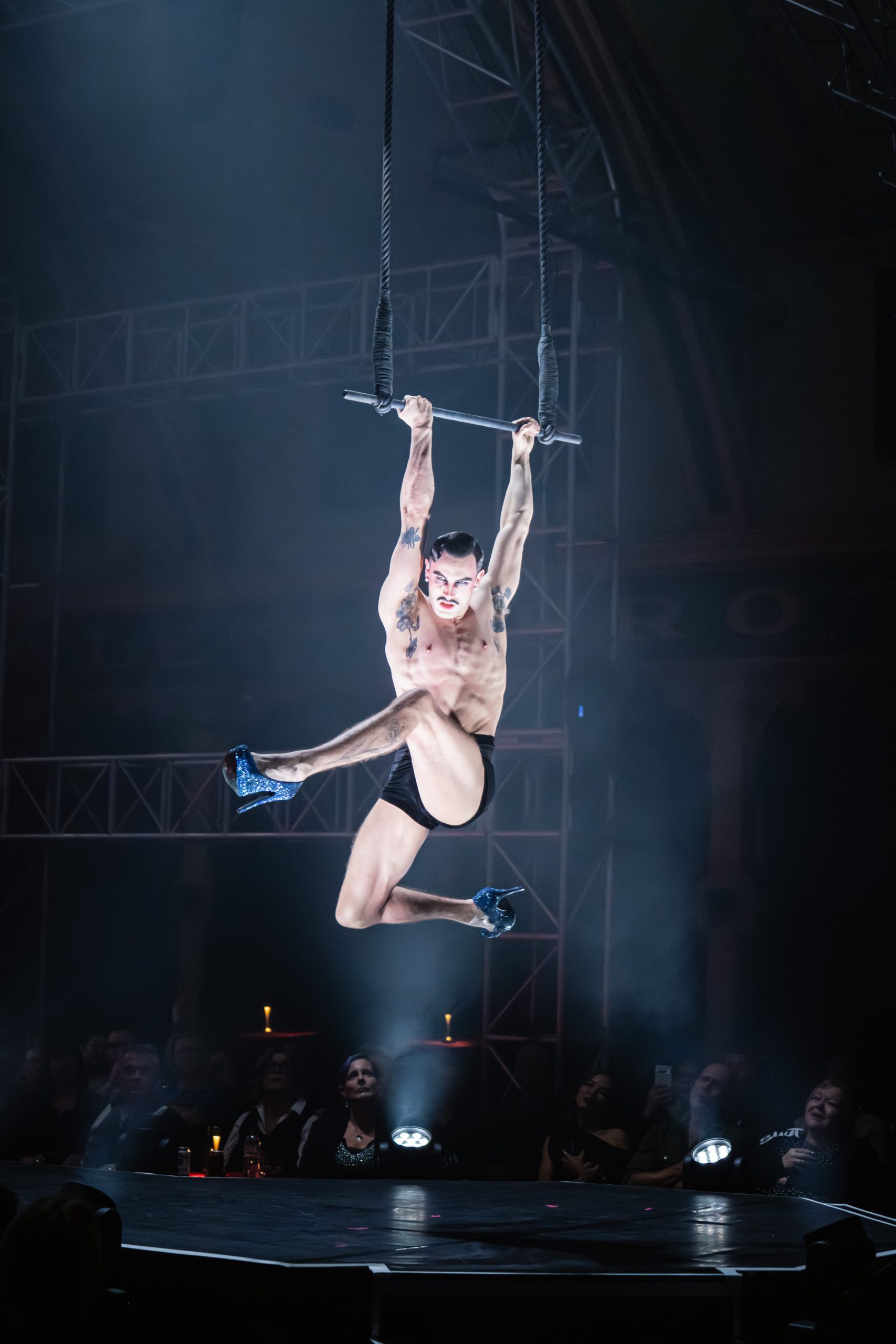 Jarred Dewey performing on aerial trapeze at Club Kabarett wearing glitter heels and black shorts under dramatic stage lighting
